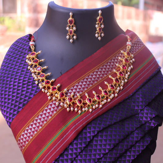 A manikratna badam shape necklace with gold plating and red gemstones, displayed on a mannequin wearing a purple and red saree, accompanied by matching earrings.